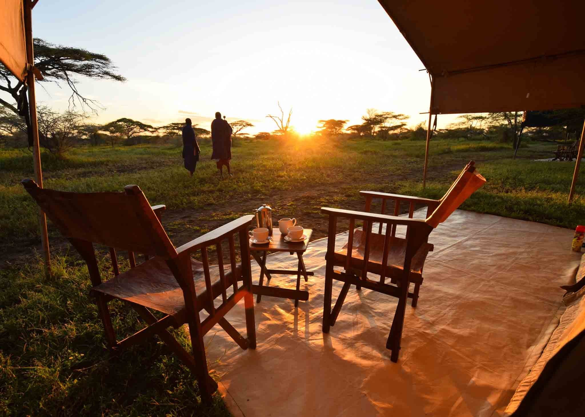 Safari chairs and a coffee tray on a tented camp veranda at sunset, with two figures walking on the plains beyond